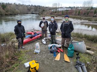 Four young men stand at the edge of the river. They are wearing slacks, sweaters and medical (COVID) masks. In front of thediving equipment like flippers, oxygen tanks, diving masks and assorted bags, are laid on the grounnd. Immediately behind them a white canoe and a red kayak laie on the ground. Beyond the canoe and the kayak lies the waters edge and then the river. In the far backgroun there are larege copses of trees and from within these, several buildings and one bridge poke through the vegetation.