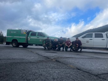 A photo of five people posing for the picture in front of a green wildland fire engine and a white truck.