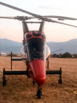 A helicopter with a needle-nosed, wedge-like profile and extruding half-sphere side windows sits on a grassy field with mountains behind.