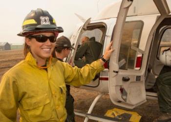 A female firefighter in uniform smiles while holding open the door to a helicopter