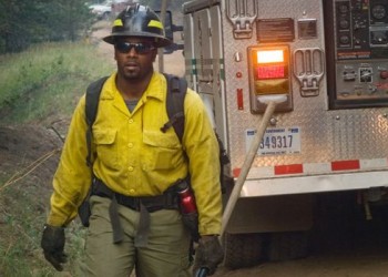 A firefighter in uniform carrying an axe walks in front of a firetruck