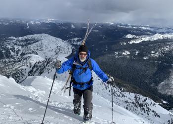 A man hikes in deep snow with skis on his back