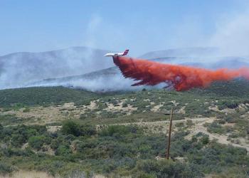 Air tanker dropping orange retardant near houses with smoke in the background.