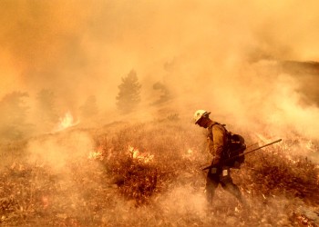 A firefighter carries a tool while walking through flames and smoke to add fireline