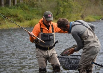 Two men fish on a river, one pulling one large fish into a net, while the other holds a rod.