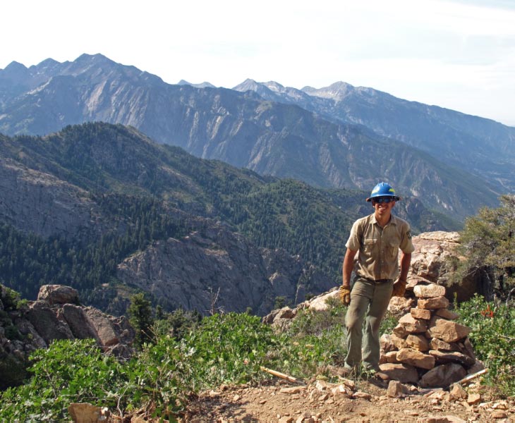 An employee with the Forest Service Wilderness crew stands on a trail with a scenic mountain range.