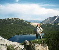 Photo of a man standing on a boulder, below and in the background is a lake.