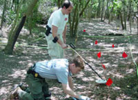 Two men, one on his knees, search a forest floor with a metal detector.