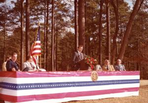 US Senator Dale Bumpers giving a speech at the rededication of the Crossett Experimental Forest.  From left to right:  Larry E. Lassen, Director, Southern Forest Experiment Station (obscured); J.E. Martin, University of Arkansas Vice President; Robert E. Buckman, Deputy Chief for Research, USDA Forest Service; Don Gammel, CEF forest manager (standing, behind US flag); Senator Bumpers; Reverend Herman Reese, St. John's Lutheran Church in Crossett;  John Wishart, Georgia-Pacific (obscured); B.G. Gresham, Arkansas State Forester; and Russell R. Reynolds, USDA Forest Service (retired).