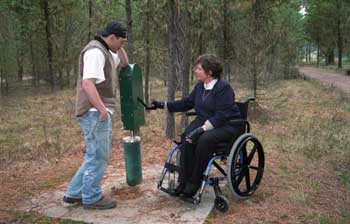 Photo of two people at a handpump in a forested area. A woman using a wheelchair is operating the accessible pump with one hand, while the young man watches.