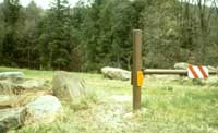 Photo of a trail going around a road closure gate. The space between the gate end post on the right and a large boulder on the left is sufficient for passage by a person using a wheelchair, but not a vehicle.