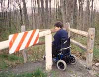 Photo of a woman using a wheelchair maneuvering through a V-shaped rail fence gate bypass.