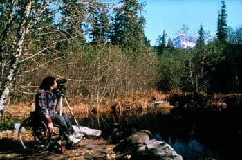 Photo of a man using a wheelchair beside a lake. He is looking through a tripod-mounted camera with a telephoto lens at a snow-covered mountain in the background.