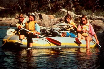 Photo of four people rafting on a river. The raft contains camping equipment and a wheelchair.
