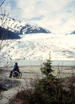 Photo of a person in a wheelchair on a paved path looking across a lake at a glacier.
