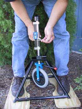 Photo of a person adjusting the pressure on the wheel of a rotational penetrometer. The rotational penetrometer is a wheelchair caster that is mounted within a frame. The frame holds the caster in position against the ground while it is swiveled by hand using an attached rod. This action simulates wheelchair traffic on a surface.
