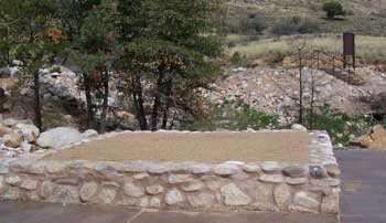Photo of a raised tent platform adjacent to a hill on one side and asphalt paved outdoor recreation access route on two sides. The platform is constructed of mortared native rock and is filled with fine gravel.