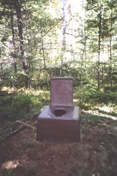 Photo of a pit toilet with no walls, surrounded by forest. The toilet is a simple square riser with an integral seat and a backrest lid.