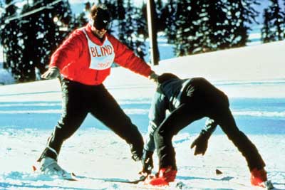 Photo of two people downhill skiing. One person is an instructor and is skiing backwards. The other person is wearing a vest that reads: BLIND.