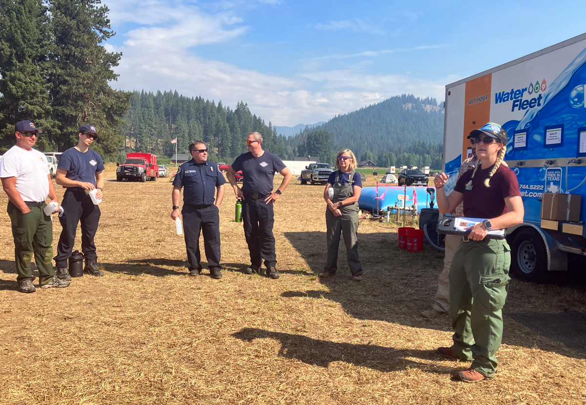 "A group of people in a forest meadow standing next to a Water Fleet truck listening to another person speaking."