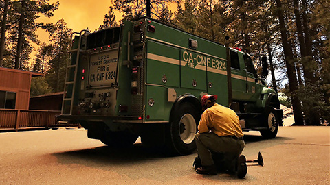 A green fire truck with a firefighter dressed in yellow Nomex shirt and a hard hat kneeling next to it with orange wildfire clouds behind it.
