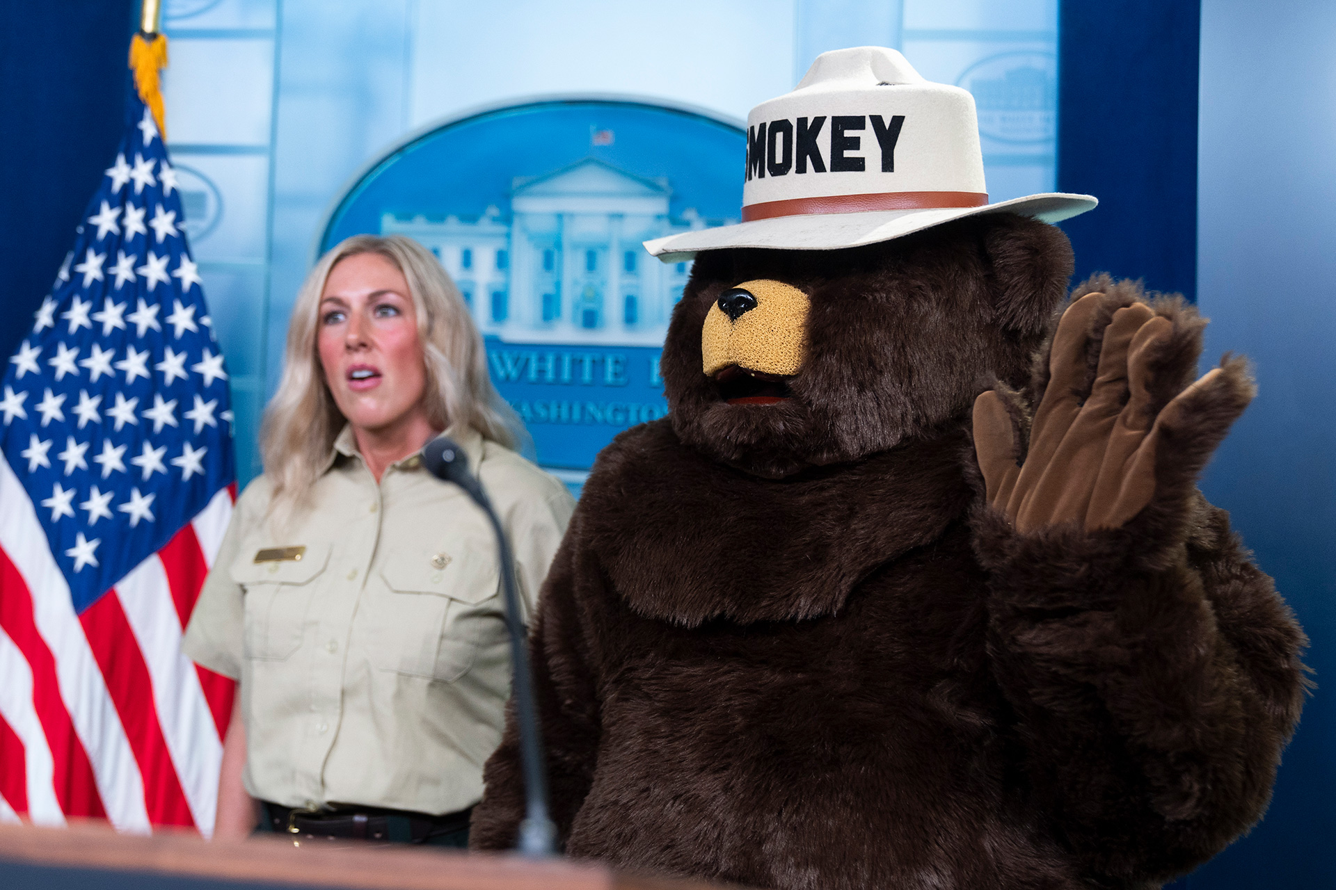 Smokey Bear and a USDA Forest Service employee in uniform standing at the podium in the White House Press Briefing room. An American flag appears on the left side, and the White House logo appears on the wall behind.