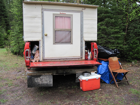 A truck camper and vehicle parked in the forest with a food cooler and used dishes sitting in the open, demonstrating improper food storage.
