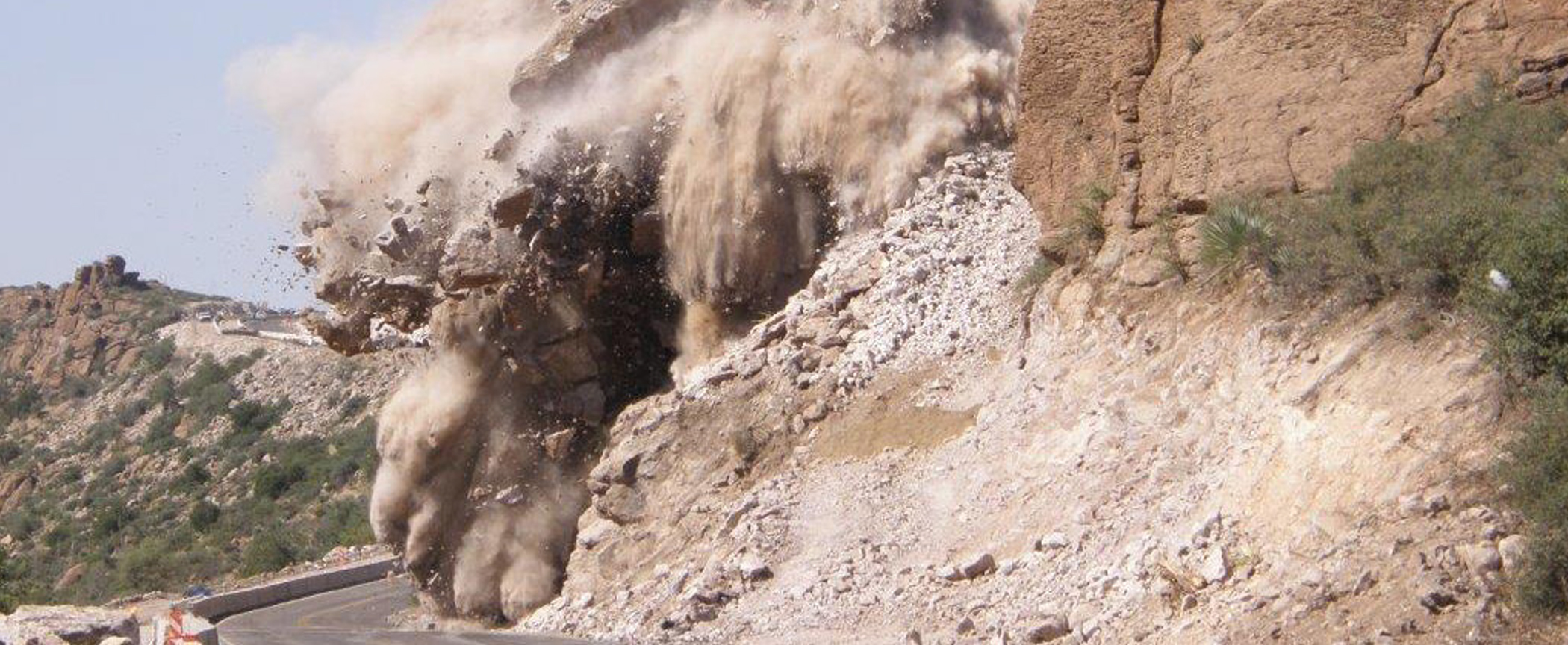 Photo: cloud of debris and dirt blown from side of mountain scatters across road after blast.