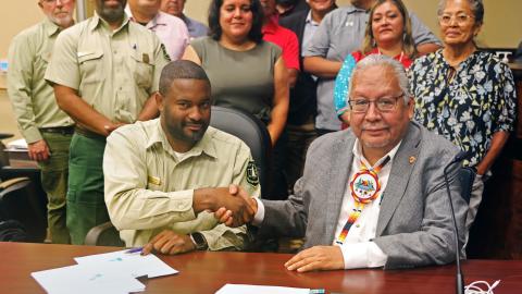 Photo of Forest Service employees shaking hands with a Tribal leader wearing a business coat wearing a beaded medallion sitting down at a table with signed agreements.
