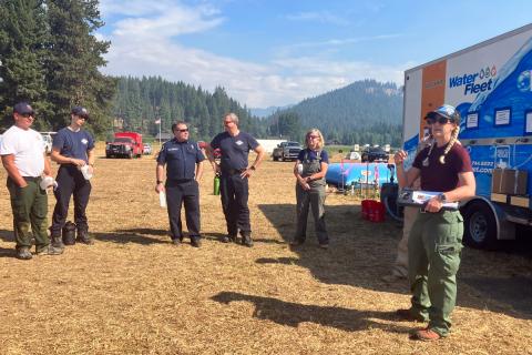 A group of people in a forest meadow standing next to a Water Fleet truck listening to another person speaking.