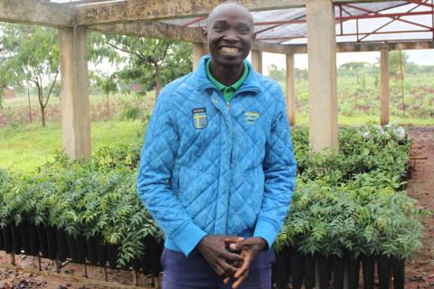 A man stands smiling in front of hundreds of tree seedling in rows of small pots.