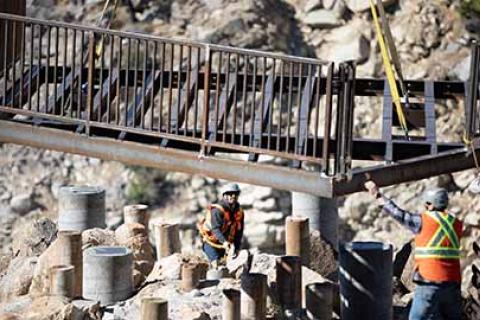 Forest Service contractors move a bridge onto pillars through a canyon using a crane.