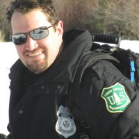 A picture of Matt Jemmett, wearing sunglasses, with a jacket displaying the U.S. Forest Service insignia and law enforcement badge.
