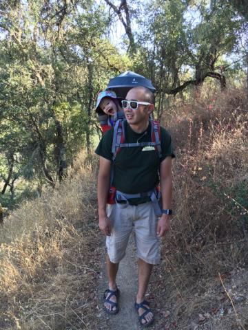 A mak in khaki shorts and black t-shirt, strolls through the woods with a smiling baby in a baby carrier on his back.
