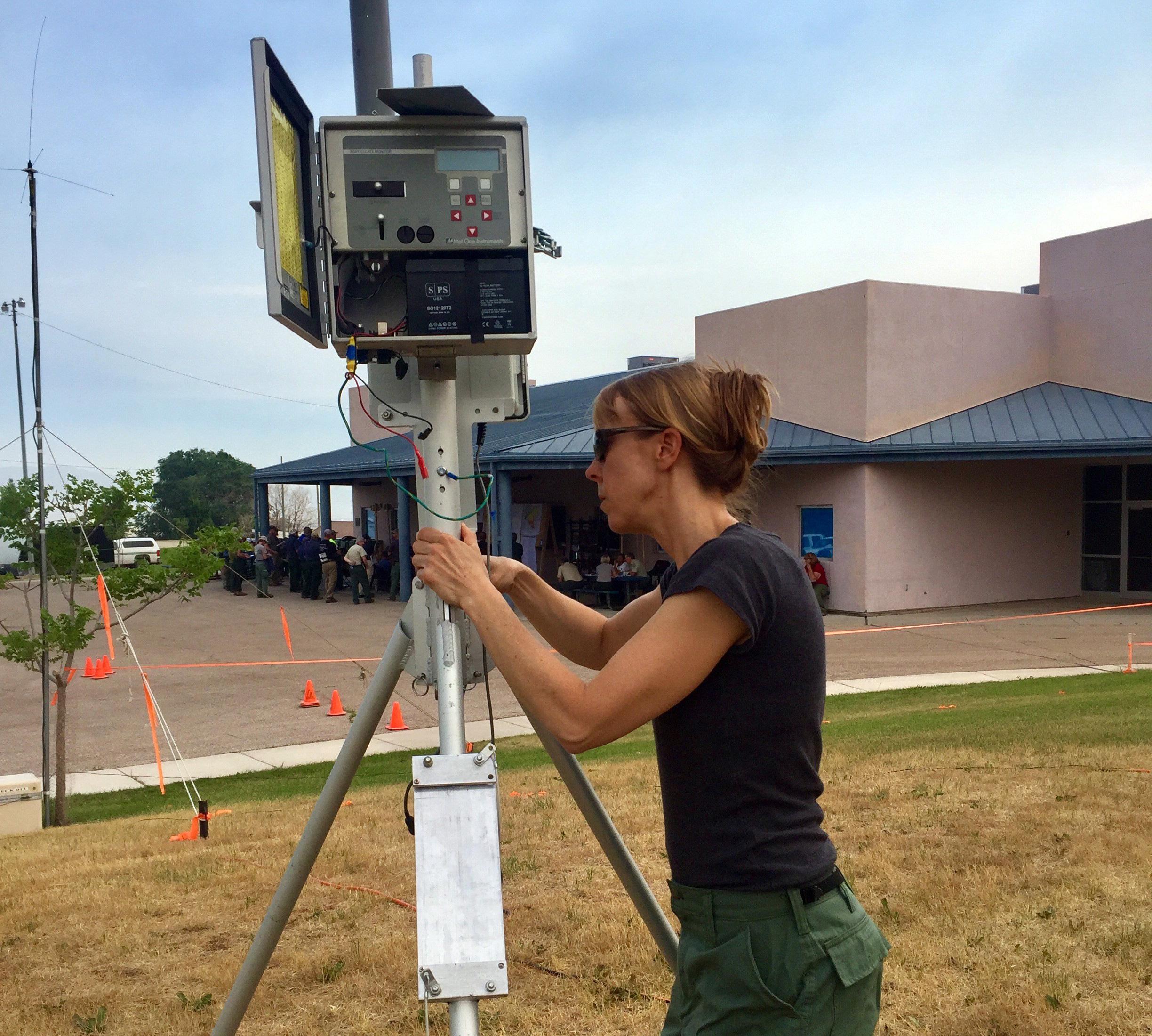 Woman with hair tied back sets up small, square electronic box atop a metal tripod. 