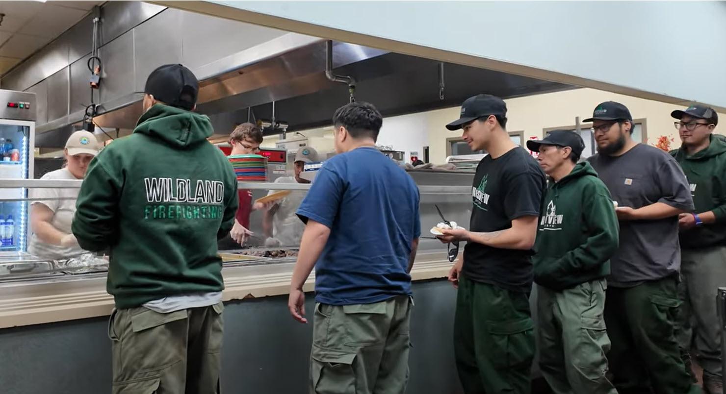 Image shows wildland firefighters standing in line in a cafeteria to get food.
