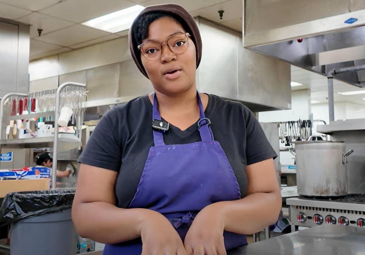 Image shows a young woman in an apron and hairnet addressing the photographer while standing in a cafeteria kitchen.
