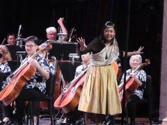 Woman standing on stage with a symphony leads the audience in a hula.