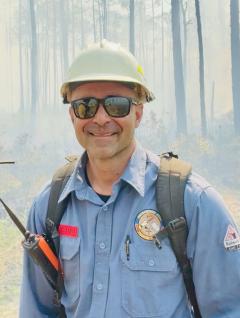 Photo of a man (from the chest up, wearing a blue shirt, backpack, walkie talkie clipped on the right strap, a white hard hat and protective sunglasses. In the background a forest engulfed in smoke.