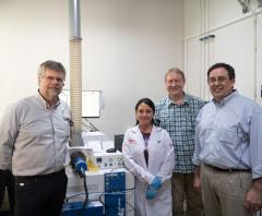 Four people stand to either side of machinery. Three men and one woman; she is wearing a lab coat and gloves.
