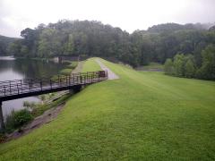 A grass covered compacted earth dam and foot bridge on a lake.