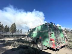 A Forest Service fire engine staged on a forest fire while smoke billows into the sky in front.