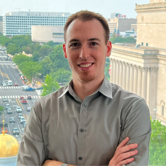 Michael Pope, wearing a collared shirt, stands with arms crossed in front of a window. Below and behind him are city buildings and traffic on the street.