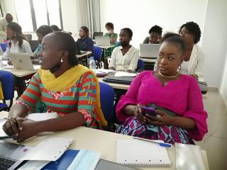 Two women in the foreground sit at a desk listening to a presentation out of the frame, one take notes in a notebook and the other on her phone. Several women in the background also listen to the presentation and take notes.
