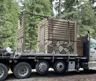 Above-ground wall unit of vault toilet tied down on semi-trailer bed.