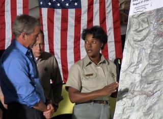 Leslie Weldon, in Forest Service field uniform, points to a large map while President George W. Bush looks on. A large American flag hangs vertically behind them. 