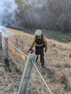 A man in FS uniform and safety gear, walking by a rustic fence, igniting a control fire with the red can in his hand.