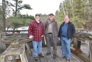 Three older men standing on a bridge, behind them a river can be seen, at each side (in the background) tall trees ris.