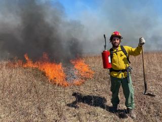 A young man in firifighting gear holding an axe upside-down by the handle with his left hand, and cradling a small, red tank with a nozzle with his right hand. He stands in a field of dried vegetation and a small prescribed fire burns in the background behind him.