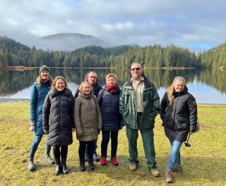 Six visitors and one Forest Service employee pause with Ward Lake and mountains of Tongass National Forest behind them. The visitors are wearing hiking shoes or boots and winter coats. The employee is in uniform and hiking boots. Bright blue sky is above them and is filled with fluffy white clouds.